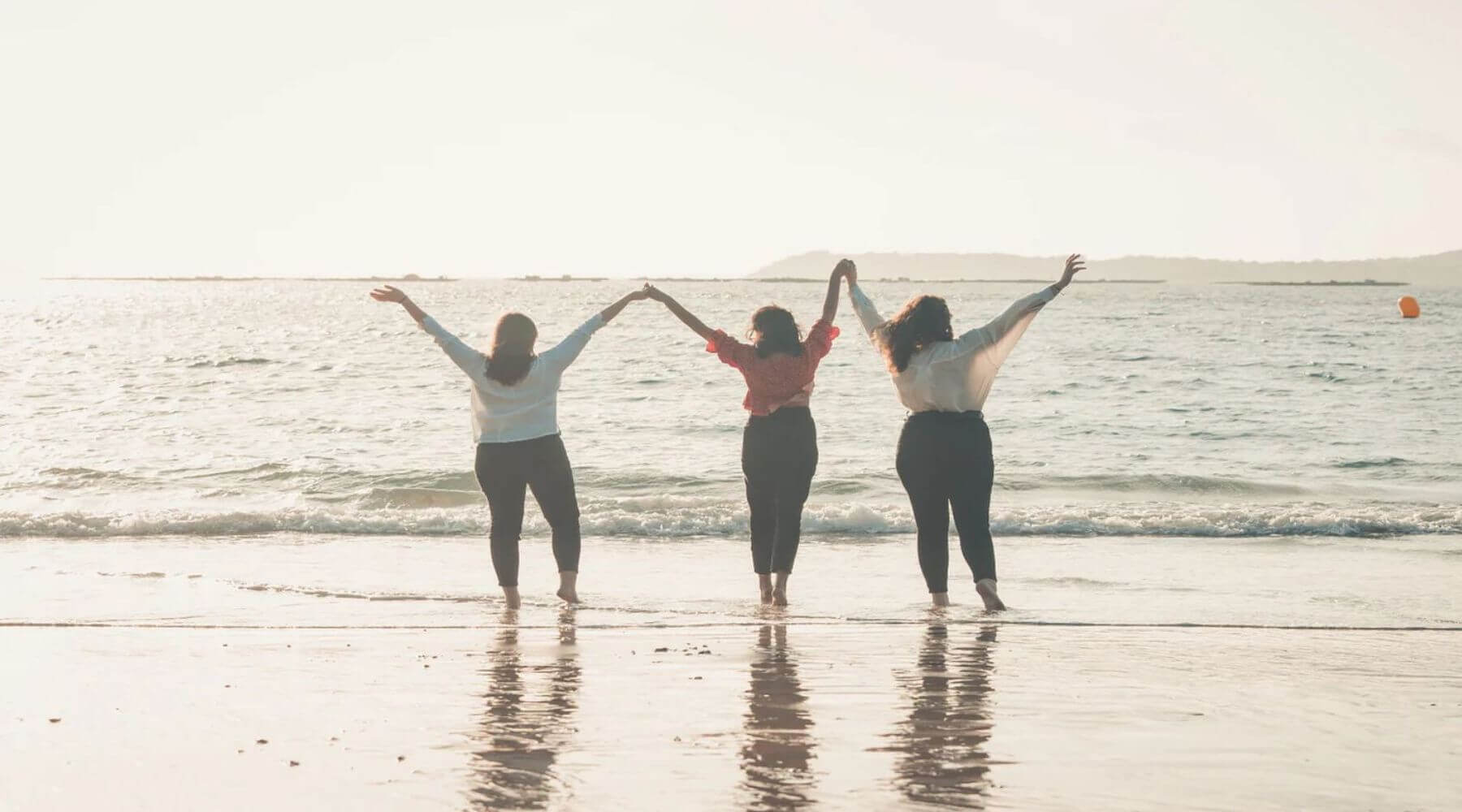 Three women on a beach with their hands raised to the sky